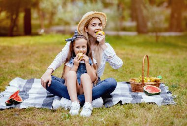 Aile pikniği. Mutlu Anne ve Küçük Kız Yeşil Park 'ta ekose üzerinde oturan ve kameraya gülümseyen elma yiyor.