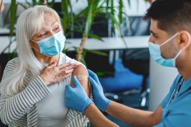 Koruyucu yüz maskeli doktora yakın çekim, yaşlı bayana mevsimsel grip veya enfeksiyon aşısı enjekte edildikten sonra hastaların kollarına bant yapıştırıyor. Sağlık ve Sağlık Hizmetleri