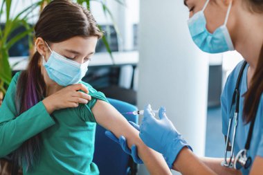 Küresel salgın sırasında yeni modern grip aşısı için klinik deney. Hastanede yüz maskesi takan küçük bir kız iğne oluyor. Doktor ya da hemşire şırıngayla kadın hastaya iğne yapıyor.