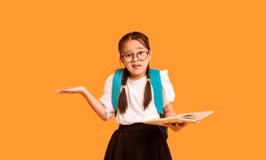 Confused Korean Schoolgirl Holding Book Shrugging Shoulders Posing Over Yellow Studio Background. Back To School Concept. clipart