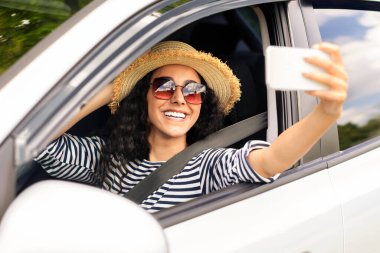 Güneş gözlüklü, yaz şapkalı, koca ya da erkek arkadaşıyla araba yolculuğunun keyfini çıkaran, elini araba camından uzatan ve akıllı telefon kullanarak selfie çeken orta doğulu güzel bir kadın.