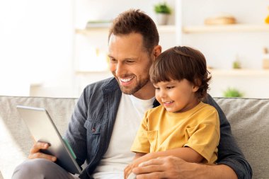 Modern teknolojiler ve çocuk gelişimi. Mutlu genç baba dijital tablette çizgi film izliyor, evde birlikte oturuyor, boş yer var.