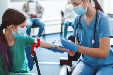 Hemşire küçük hastaların damarına iğne batırıyor. Kolunu tıbbi turnikeyle tutuyor. Kan testi için hazırlık. Tıbbi üniformalı ve maske takan kadın doktor örnek topluyor. Sağlık Hizmetleri İlacı