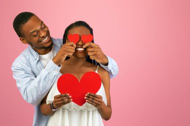 Aşk Konsepti Gözümü kör etti. Sevecen Afrikalı Amerikalı çift, ellerinde kırmızı kağıt kalp, kız arkadaşlarının gözlerini kapatan gülümseyen adam, pembe stüdyoda eğlenen romantik erkek ve kadın.