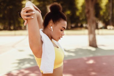 Kollar için egzersiz, dışarıda spor. Bin yıllık sarı sutyen, havlu ve kablosuz kulaklık takan siyahi kadın profili halka açık parkta elini kolunu sallayarak dolaşıyor. Eğitimdeki model sporcu