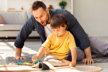 Arkadaş canlısı aile kavramı. Küçük şirin çocuk babasıyla ahşap tren oyunu oynuyor. Şaşırmış adam heyecanla oyuncağa bakıyor, evde yerde oturuyor.
