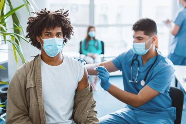 Doktor ya da hemşire bir hastanın omzuna aşı ya da aşı yapıyor. Küresel salgın sırasında kitle aşılama ve grip veya korona virüsüne karşı koruma. Afrikalı maskeli adam kameraya bakıyor.