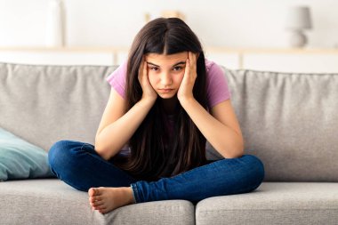 Sinirli Hintli genç kız kanepede oturuyor, sıkılmış, iletişim eksikliği çekiyor, Coronavirus tecridi sırasında evde kapana kısılmış, gençlik depresyonu geçiriyor, tam boy