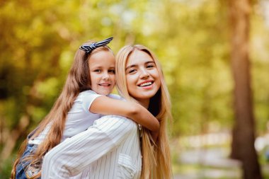 Anne, küçük kızını sırtında taşıyor. Dışarıda park yerinde kameraya gülümsüyor. Aile Hafta sonu Konsepti