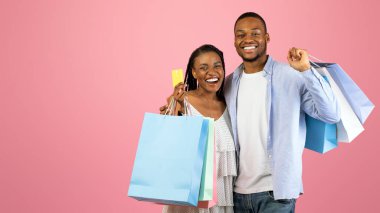 Tüketim ve İlişki Konsepti. Gülümseyen siyah bir çiftin portresi kredi kartı ve alışveriş çantaları gösteriyor, pembe stüdyo arka planında tek başlarına duruyorlar.