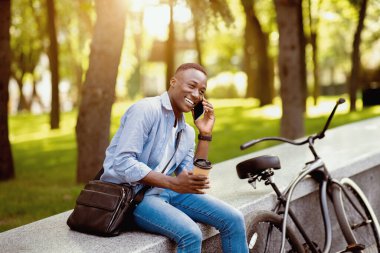 Bisikletli ve kahveli kendinden emin Afro-Amerikalı adam şehir parkında cep telefonuyla konuşuyor.