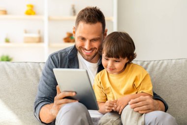 Çocuk gelişimi içeriği. Genç, pozitif baba, sevimli oğluna dijital tablet gösteriyor. Çocuk internette eğitim programından zevk alıyor. Evdeki kanepede oturuyor.