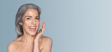 Bir kadın yüzüne neşeli bir ifadeyle cilt ürünleri sürüyor. Sakin, minimalist bir çevrede kendine özen ve güzelliği yumuşak renklerle kucaklıyor..