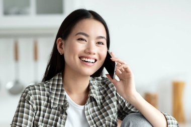 Genç Asyalı bayan sağlıklı kahvaltı yapıyor ve telefonda konuşuyor, gülümsüyor ve fotokopi odasına bakıyor. Neşeli kadın mutfakta oturuyor, yulaf ezmesi yiyor ve kız arkadaşı ya da sevgilisiyle sohbet ediyor.