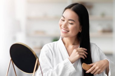 Bornozlu neşeli genç Asyalı bayan yatak odasında oturuyor, ahşap fırçayla güzel ipek saçlarını tarıyor, aynaya bakıyor ve gülümsüyor, kopya odası. Saç bakım ürünleri kavramı