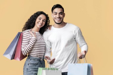 Sarı arka planda alışveriş torbaları olan neşeli, genç Orta Doğulu çift pozları, Alış-veriş yapan mutlu bir Arap ve kadın, sezonluk satışların tadını çıkarıyor, kopyalama alanı.