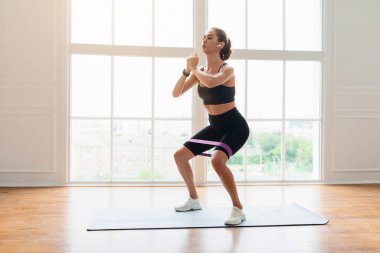 Eğitim Konsepti. Kendinden emin sporcu kadın fitness stüdyosunda elastik direnç bandıyla ya da oturma odasında bacak ve kalça egzersizi yaparak kulaklıkla müzik dinleyerek çömeliyor.