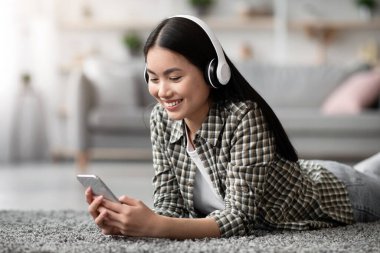 Oturma odasında kablosuz kulaklığı olan güzel Asyalı bayan yepyeni cep telefonu kullanıyor, internetten film veya video izliyor, hafta sonunun keyfini çıkarıyor, evi kopyalıyor.