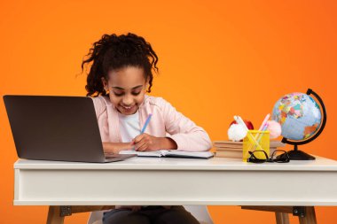 Uzaktan eğitim konsepti. Gülümseyen genç zenci kız masada oturuyor, dizüstü bilgisayar kullanıyor ve defterine ya da günlüğüne bir şeyler yazıyor. Afrikalı liseli kız çevrimiçi ders izliyor, not alıyor, ödev yapıyor.