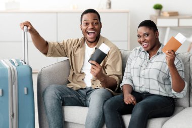 Evdeki koltukta oturan ve kameraya gülümseyen bavullu mutlu siyah aile çift seyahat biletleri taşıyor. Tatilinizi Kutlayan Gezginler 'iniz kutlu olsun. Seyahat ve Turizm Konsepti