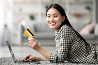 Mutlu genç Asyalı kadın oturma odasında halıya uzanıyor ve dizüstü bilgisayar ve kredi kartı kullanıyor, yeni web sitesinde online alışveriş ya da bankacılık, ev içi, fotokopi alanı, yan görüş