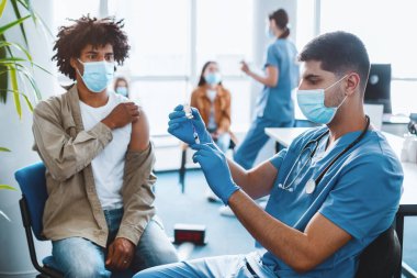 Coronavirus bağışıklığı. Şırınganın şırıngasından şırınga aşısı alan mavi lateks eldivenli ve koruyucu maskeli hasta arka planda bekliyor.