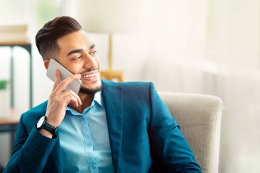 İş elbisesi içinde neşeli bir Arap girişimci ofiste cep telefonuyla konuşuyor, koltukta oturuyor, arkadaşı veya müşterisiyle sohbet ediyor. Resmi ofis çalışanı kapalı alanda akıllı telefondan konuşarak giyer.