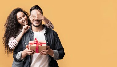 Sürpriz Hediye. Romantik genç Arap kadın, erkek arkadaşının gözlerini kapatıyor ve hediye veriyor. Mutlu bayan, sevgilisini şaşırtırken sessizlik işareti gösteriyor. Sarı arka planda birlikte duruyor.