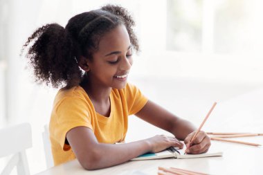 Hoş bütünlüklü Afro-Amerikan genç kız, günlük ev ödevlerini yapıyor, rahat mutfağında oturuyor, not alıyor ya da plan yapıyor, kalem ve not defteri kullanıyor, kopyalama alanı, yan görüş kullanıyor.