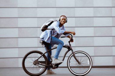Sırt çantalı ve kulaklıklı neşeli siyah adam bisiklet sürüyor. Tuğla duvarın yanında.