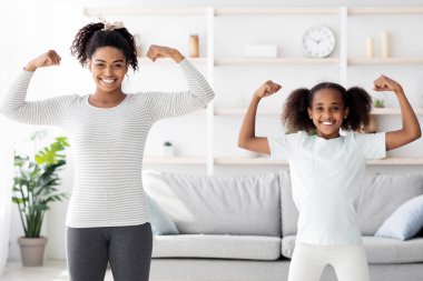 Spor giyim bölümünde güçlü Afro-Amerikan anne ve genç kız sabah egzersizlerini birlikte yaparken kaslarını gösteriyorlar, atletik siyah aile pazılarını gösteriyor ve gülümsüyor.
