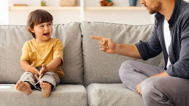 Aile içi şiddet ve çocuk istismarı. Öfkeli baba, küçük oğluna kızmalı, ağlayan çocuk, gücenmiş hissediyor, evde kanepede oturuyor.