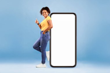 Beyaz ekranlı akıllı telefonun yanında duran güzel bir Afro-Amerikalı kadın. Mavi arka planda el hareketleriyle işaret ediyor. Genç bir kadın yeni mobil uygulama, reklam sitesi, boş alan öneriyor.