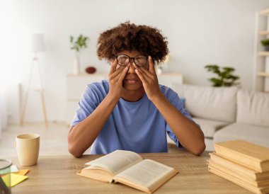 Yorgun gözlerini ovuşturan siyah bir genç, açık ders kitaplarıyla masada oturuyor, evde öğrenim görmekten yorulmuş. Uzaktan öğrenme yorgunluğu çeken Afro ergen, kitaba çok uzun bakıyor.