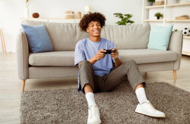 Playstation 'da video oyunundan hoşlanan mutlu siyahi genç, evde uzun süre eğleniyor. Joy Afro-Amerikan ergen oyuncu elinde joystickle yerde oturmuş bilgisayar oyunu oynuyor.