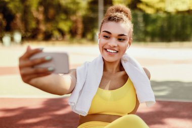 Gülümseyen siyah kadın spor molasında akıllı telefondan selfie çekiyor, kablosuz kulaklık takıyor, spor sutyen ve havlu takıyor, fitness eğitmeniyle online konferans yapıyor veya video blogu kaydediyor.