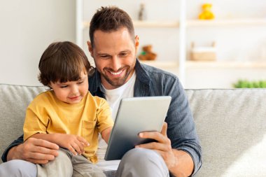 Çocuklar için eğitim uygulaması. Sevimli küçük çocuk dijital tablette video oyunları oynuyor, babasıyla koltukta oturuyor, boş yer var.
