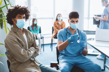 Aşı ve Bağışıklık Kampanyası kavramı. Doktor, hastaya iğne yapmadan önce aşıyı şişeden çıkarıyor. Tıbbi maskeli bir adam klinikte veya hastanede grip aşısı olmayı bekliyor.