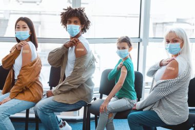 Aşılanmış çok ırklı maskeli insanlar, aşı olduktan sonra omuzlarını yapıştırıcı bandajla gösteriyorlar, klinikte sırada bekliyorlar. Yara bandı olan kadın ve erkeklerden oluşan bir grup