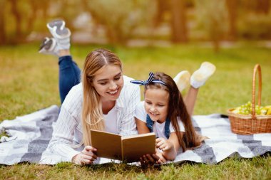 Anne ve küçük kızı açık havada ekose üzerinde uzanmış piknik kitabı okuyorlar. Yaz Hafta Sonu Konsepti