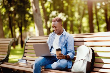 Uzaktan eğitim konsepti. Sırt çantası, kitap ve dizüstü bilgisayarı olan havalı siyah öğrenci parkta online derse katılıyor. Milenyum Afrikalı Amerikalı adam dışarıda web konferansına katılıyor.