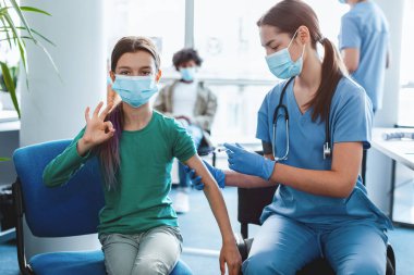 Başarılı grip aşısı yapıldı. Yüzü maskeli ergen bir kız aşıyı omzuna enjekte ederken ve klinik geçmişinde poz verirken iyi bir işaret hareketi gösteriyor. Dokunulmazlık Onaylanıyor