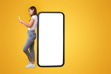 Harika bir uygulama. Beyaz ekranlı ve cep telefonu kullanan mutlu bayan, sosyal medyada neşeli kadın sohbeti, turuncu arka planda durmak, görüntüyü taklit etmek, vücut uzunluğu...