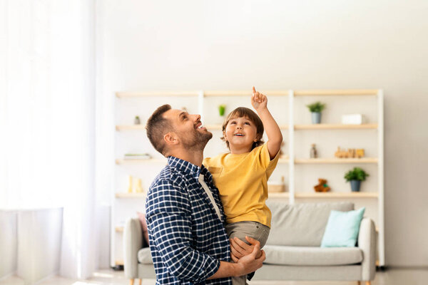 Daddy, look up. Excited little boy looking upwards with his father, child pointing with finger, man holding kid on hands, standing together at home