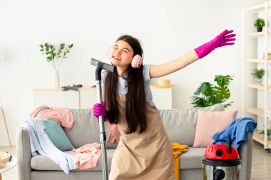 Mutlu Hintli genç kız evini temizliyor, kulaklık takıyor, elektrikli süpürgeyi mikrofon olarak kullanıyor ve eğleniyor. Mutlu ergenler ev işlerinden zevk alıyor, ev temizliği yapıyor.