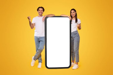 Öyle mi? Heyecanlı çift, boş beyaz ekranlı, sallanan yumruklu, neşeli adam ve kazanan kadın, sarı arka planda duran, sahte kolaj, tam vücut uzunluğu olan büyük akıllı telefona yaslanıyor.