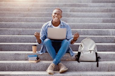 Uzaktan eğitim konsepti. Diz üstü bilgisayarı olan heyecanlı genç siyahi adam merdivenlerde EVET işareti yapıyor. Başarılı Afrikalı Amerikalı öğrenci sınavı geçtiği için mutlu, başarısını kutluyor.