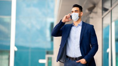 Ortadoğulu genç girişimci resmi kıyafetler içinde telefonda konuşuyor, iş görüşmesi yapıyor, maske takıyor, iş merkezi geçmişi var, fotokopi alanı, iş yeri ve salgın hastalıklı panorama