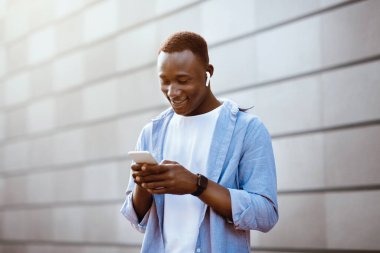 Kulaklıklı havalı siyahi adam şehirdeki tuğla duvarın yanındaki cep telefonundan müzik listesi seçiyor.