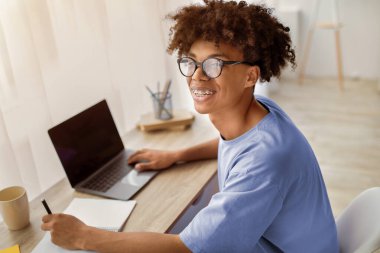 Mutlu siyah ergen dizüstü bilgisayarla masada oturuyor, uzaktan öğreniyor, evde defterine not alıyor, uzayı kopyalıyor. Neşeli Afrikalı Amerikalı genç öğrenci covid karantina sırasında çevrimiçi çalışıyor